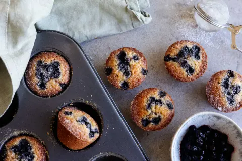 Nybakade blåbärsmuffins, en klassisk financier kaka, ligger i en form bredvid en skål med sylt och en sil.