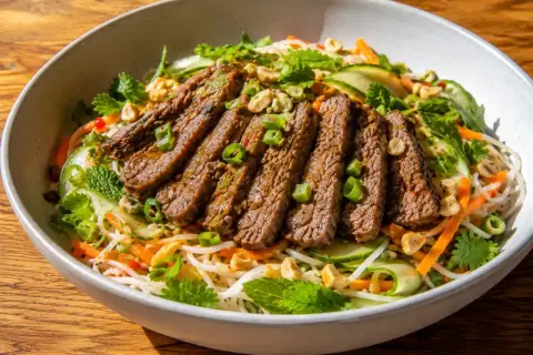 En läcker asiatisk biffsallad med tunt skivad biff, nudlar, gurka, morötter, mynta och jordnötter i en skål.