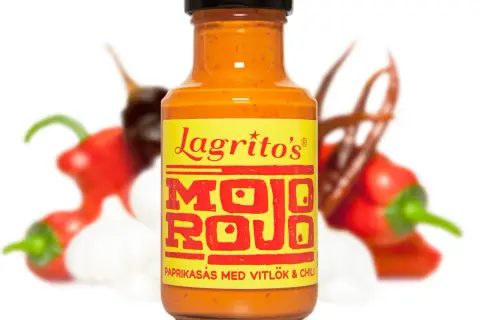 En flaska Lagrito's Mojo Rojo paprikasås med vitlök och chili, omgiven av suddiga röda chilifrukter och vitlöksklyftor.