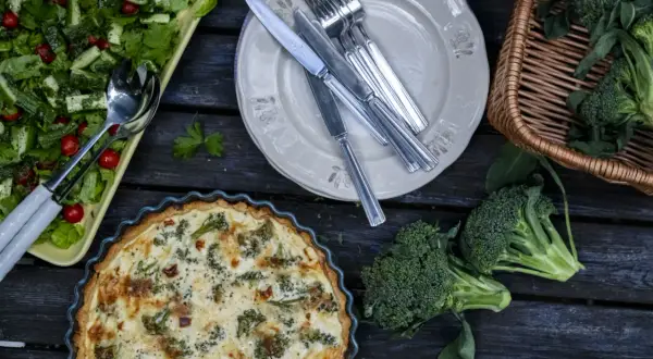 En paj med ost och broccolisås, en sallad och färsk broccoli ligger uppdukat på ett träbord.