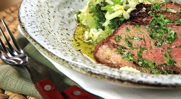 Close-up of grilled picanha steak with rice and greens, served on a speckled plate with a fork and knife.