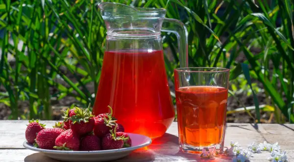 En karaff och ett glas med röd saft står på ett träbord. En tallrik med färska jordgubbar bredvid. Perfekt för ett enkelt jordgubbssaft recept.