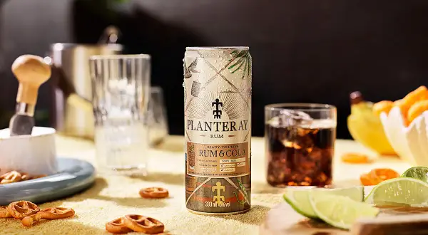 En burk Planteray Rum & Cola står på ett gult underlag, bredvid ett glas med is och mörk dryck. Små pretzels och limeklyftor ligger utspridda.