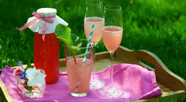 En flaska rabarberdryck och två glas med rabarberdrink med prosecco står på en bricka i gräset.
