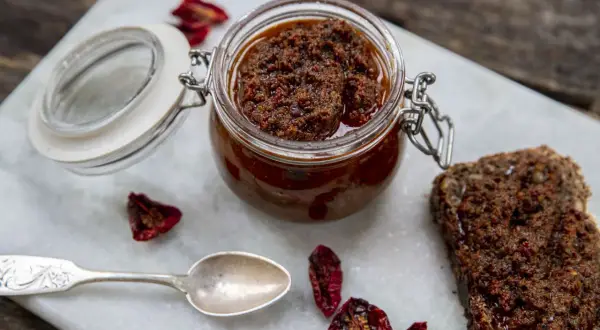 En burk med soltorkad tomat-tapenade, perfekt för ett enkelt tapenade recept. Serveras med en skiva mörkt bröd och en silver sked.