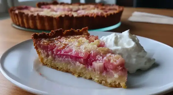 En bit glutenfri rabarberpaj med vispad grädde på en vit tallrik. Hela pajen syns i bakgrunden.