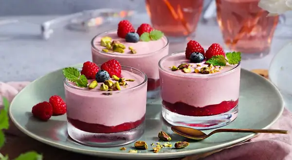 Tre glas med hallonmousse tårta, toppade med hallon, blåbär och pistagenötter. Serveras på en ljusgrön tallrik.