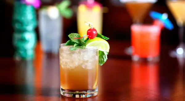 En Mai Thai-drink med is, mynta, lime och körsbär. I bakgrunden syns fler drinkar och en grön tiki-mugg.