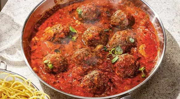 Frikadeller i tomatsås som puttrar i en panna, med en skål spaghetti vid sidan.