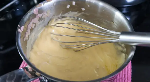 En visp rör i en gul sås i en kastrull. Små bitar av schalottenlök syns på kastrullkanten. Ett perfekt bearnaisesås recept börjar här.