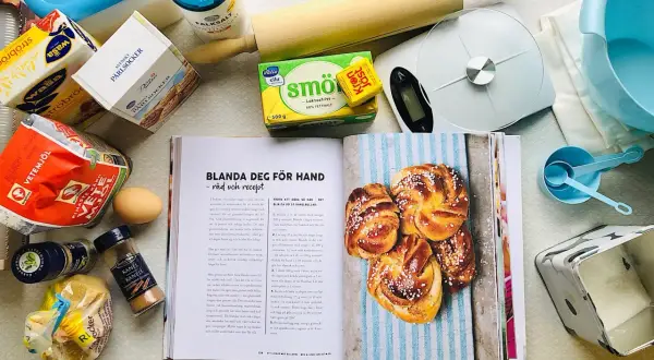 Här finns allt för ett lyckat kanelbullar recept: mjöl, jäst, smör, socker, kardemumma och kanel. En öppen bok visar hur man gör.