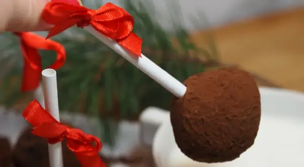 En hand håller en chokladboll på pinne med röd rosett, redo att doppas i en mugg med varm choklad.