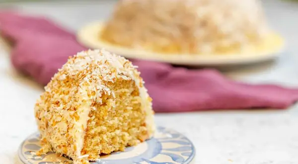 En bit sockerkaka med kokos, täckt av rostad kokosflingor. En hel kaka syns i bakgrunden.