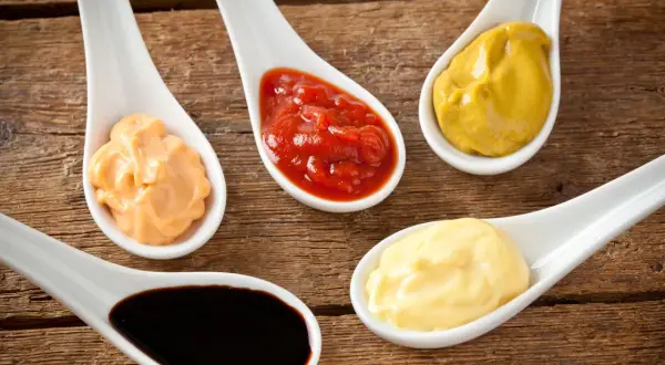 Fem skedar med olika såser: ketchup, majonnäs, senap, en orange sås och mörk bbq sås på träbakgrund.