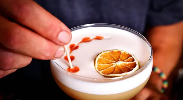 En hand dekorerar en whiskey sour med en tunn pinne, med en torkad lime och ett mönster av bitters på skummet.