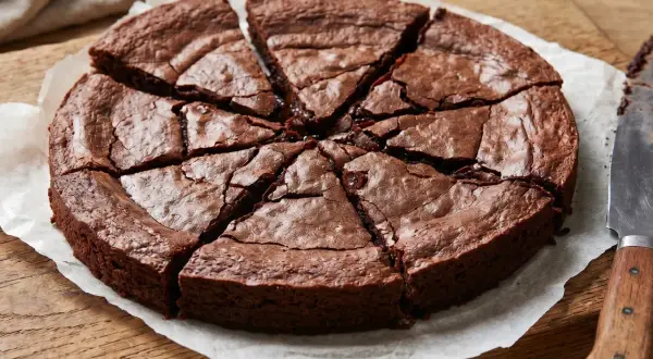 En saftig glutenfri kladdkaka, uppskuren i åtta bitar på bakplåtspapper. En kniv ligger bredvid.