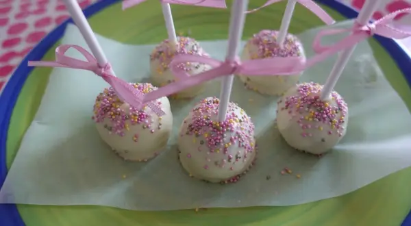 Sju vita cake pops med rosa och gula strössel, dekorerade med rosa band, ligger på ett ljusgrönt papper på en grönblå tallrik.