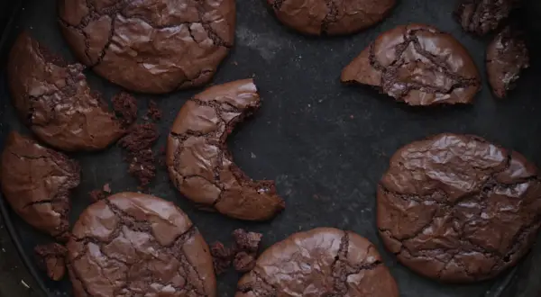 Mörka, spröda nutella kakor med sprucken yta ligger utspridda i en form. En kaka är avbruten och visar det saftiga inre.