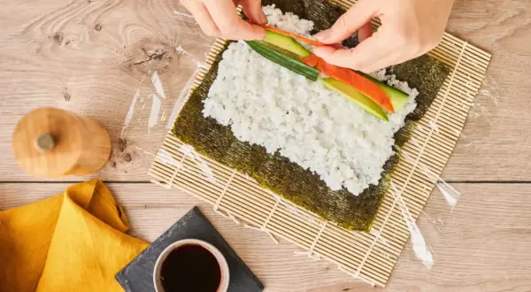 Händer lägger lax och gurka på ris för ett sushi recept.
