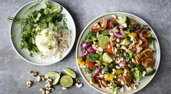 Two plates of food: one with rice, cilantro, and lime; the other with stir-fried chicken, vegetables, and cashews.