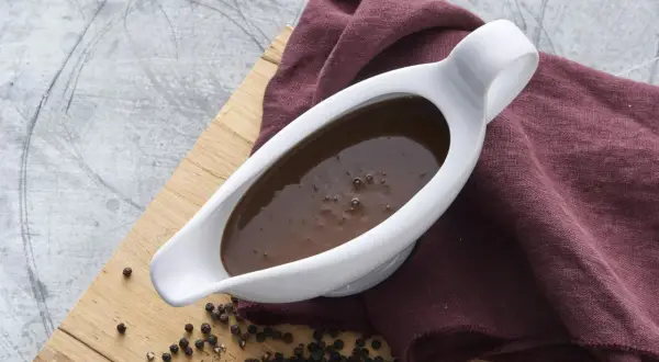 En vit såssip med mustig brunsås, redo för ett klassiskt brunsås recept. Svartpepparkorn och en vinröd duk fulländar bilden.