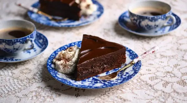 En bit kladdkaka med chokladganache och vispad grädde, serverad med kaffe på blåvita koppar.