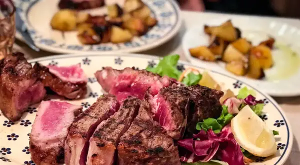 En saftig bistecca fiorentina, perfekt grillad med en rosa kärna, serveras med krispig sallad och citronklyfta. Rostade potatisar i bakgrunden.