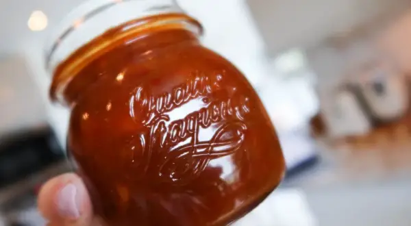 En hand håller en burk med hemlagad rabarberchutney. Den mörkröda chutneyn glänser i ljuset.