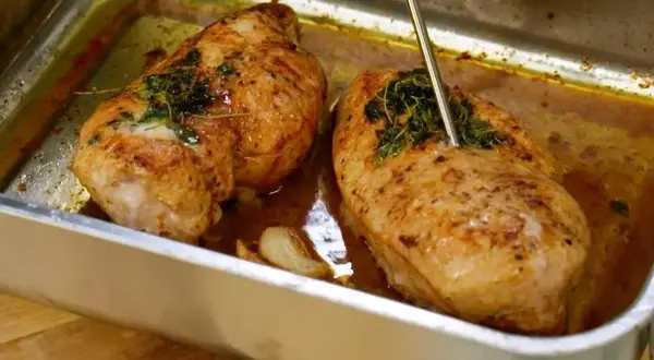 Två saftiga kycklinglårfiléer i en ugnsform, kryddade med örter och vitlök. En termometer mäter temperaturen för perfekt grillad kyckling.