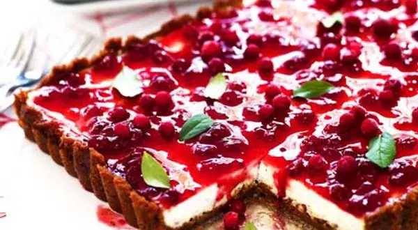 En rektangulär pepparkakscheesecake med en glansig lingonfyllning och färska lingon. Garnerad med gröna blad.