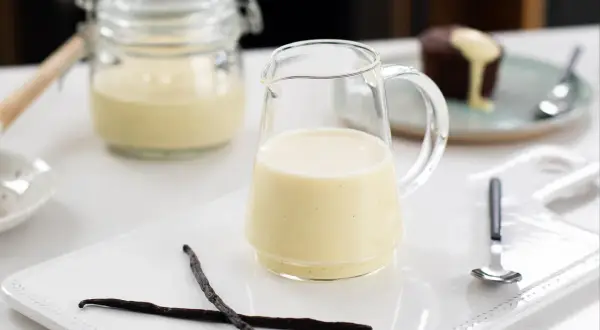 En liten kanna med krämig vaniljsås, perfekt till efterrätt. Två vaniljstänger ligger bredvid.