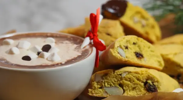 En kopp varm choklad med marshmallows och chokladbitar bredvid en hög med saffransbiscotti med mandel, inslagna i ett rött band.