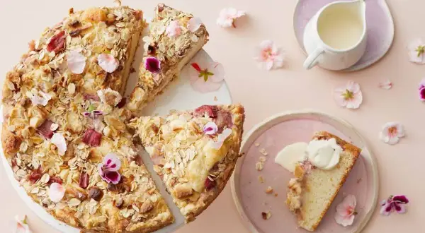 En härlig glutenfri rabarberkaka med havregryn och nötter, dekorerad med blommor. En bit serveras med grädde.