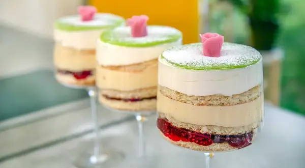 Tre vackra prinsesstårtor i glas, lager av sockerkaka, grädde och sylt, toppade med grön marsipan och en rosa ros.