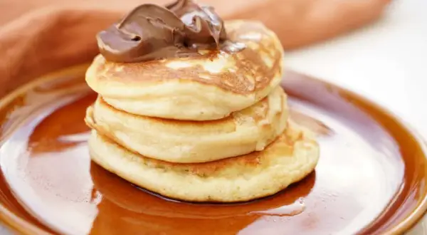 En stapel fluffiga amerikanska pannkakor toppas med chokladkräm på en brun tallrik.