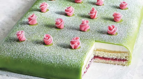 En grön täckt liten tårta med rosa marsipanrosor och pudersocker. En bit är bortskuren och visar lager av grädde och sylt.