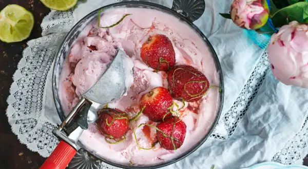 En skål med rosa jordgubbsglass, toppad med hela jordgubbar och limezest. En glasskopa vilar i skålen.