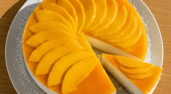 En krämig mango cheesecake med färska mangoskivor som pryder toppen. En bit är uppskuren och visar den lena fyllningen.