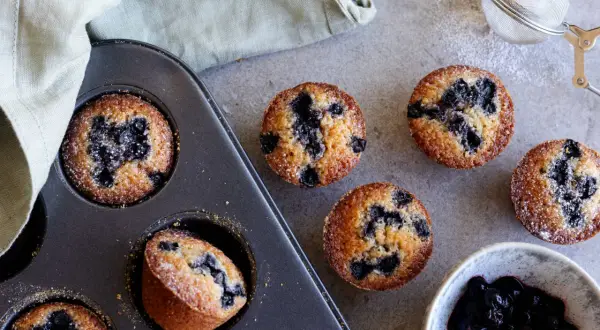 Nybakade blåbärsmuffins, en klassisk financier kaka, ligger i en form bredvid en skål med sylt och en sil.