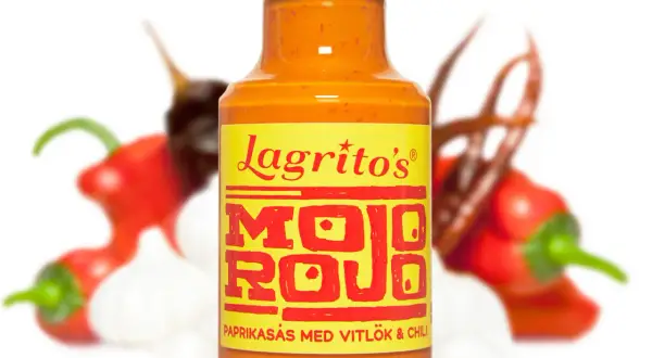 En flaska Lagrito's Mojo Rojo paprikasås med vitlök och chili, omgiven av suddiga röda chilifrukter och vitlöksklyftor.