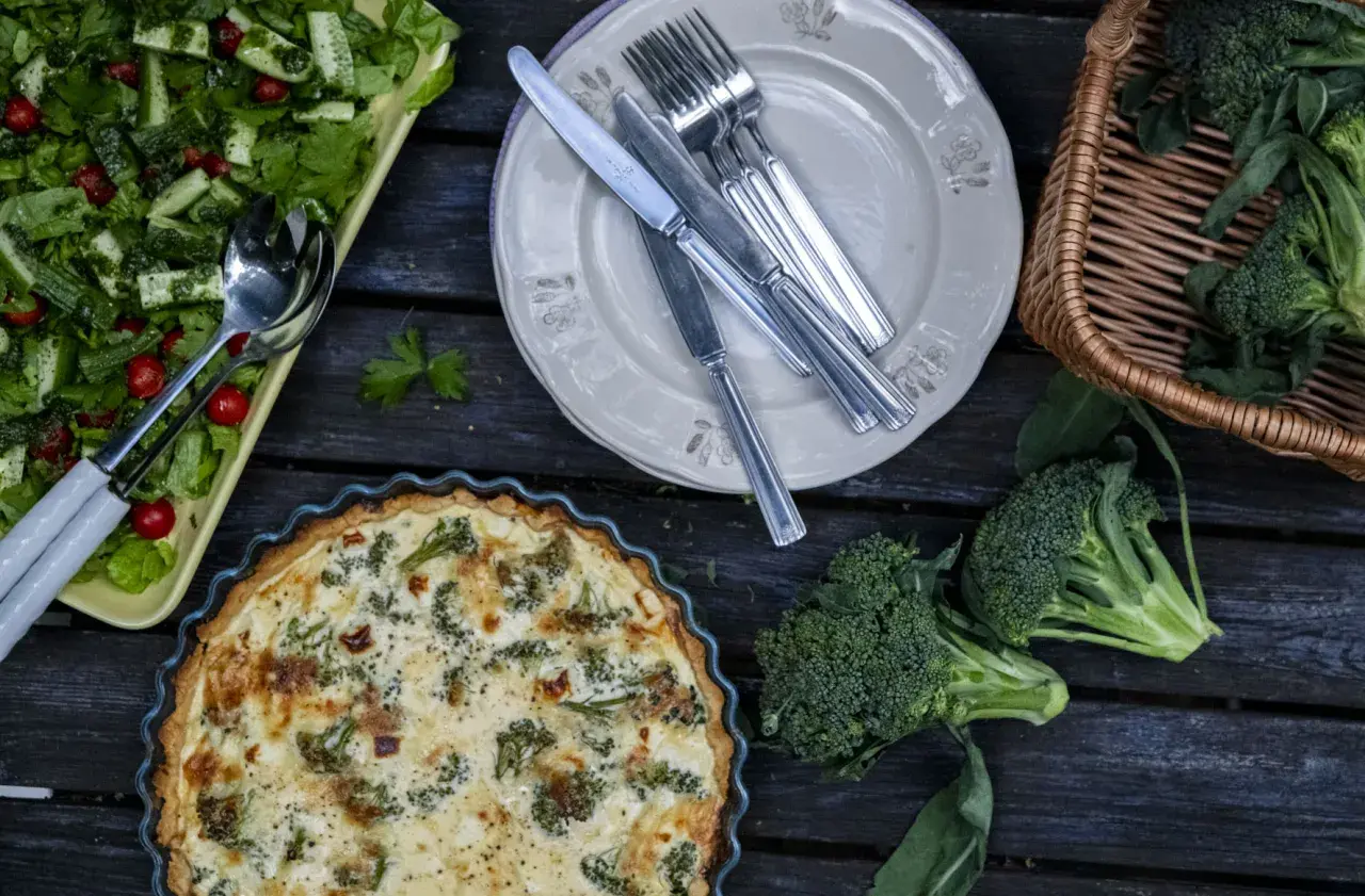 En paj med ost och broccolisås, en sallad och färsk broccoli ligger uppdukat på ett träbord.
