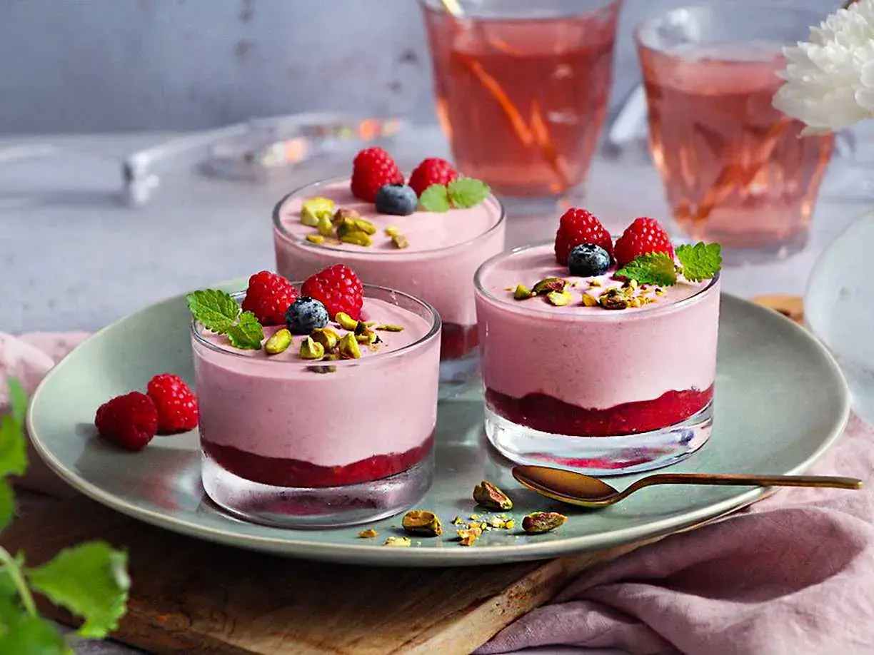 Tre glas med hallonmousse tårta, toppade med hallon, blåbär och pistagenötter. Serveras på en ljusgrön tallrik.