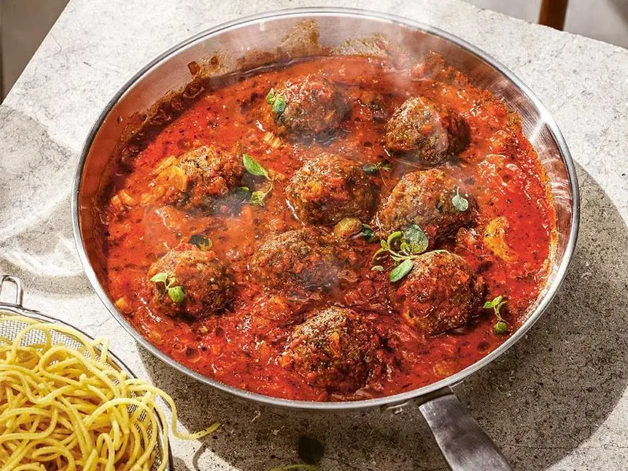 Frikadeller i tomatsås som puttrar i en panna, med en skål spaghetti vid sidan.