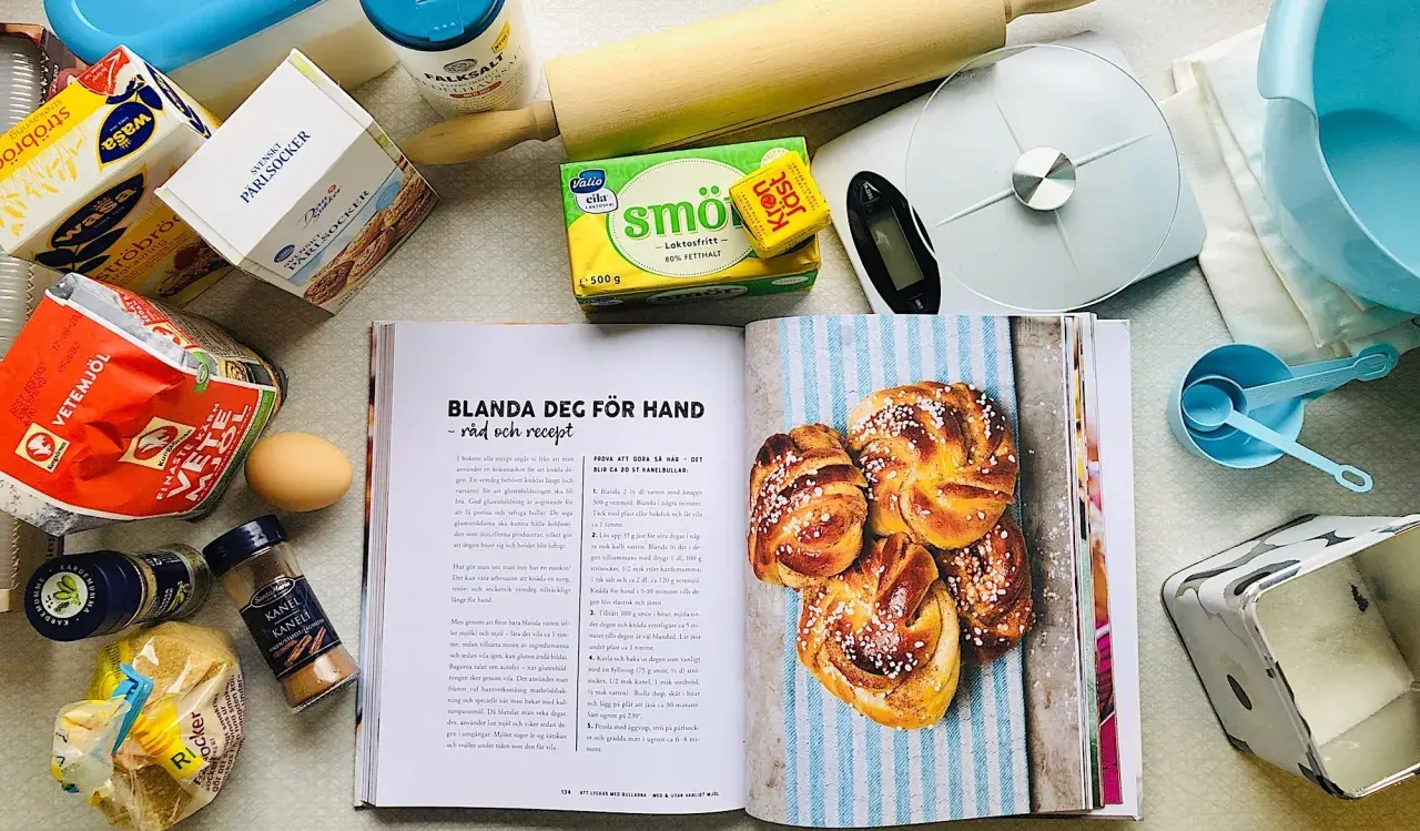 Här finns allt för ett lyckat kanelbullar recept: mjöl, jäst, smör, socker, kardemumma och kanel. En öppen bok visar hur man gör.
