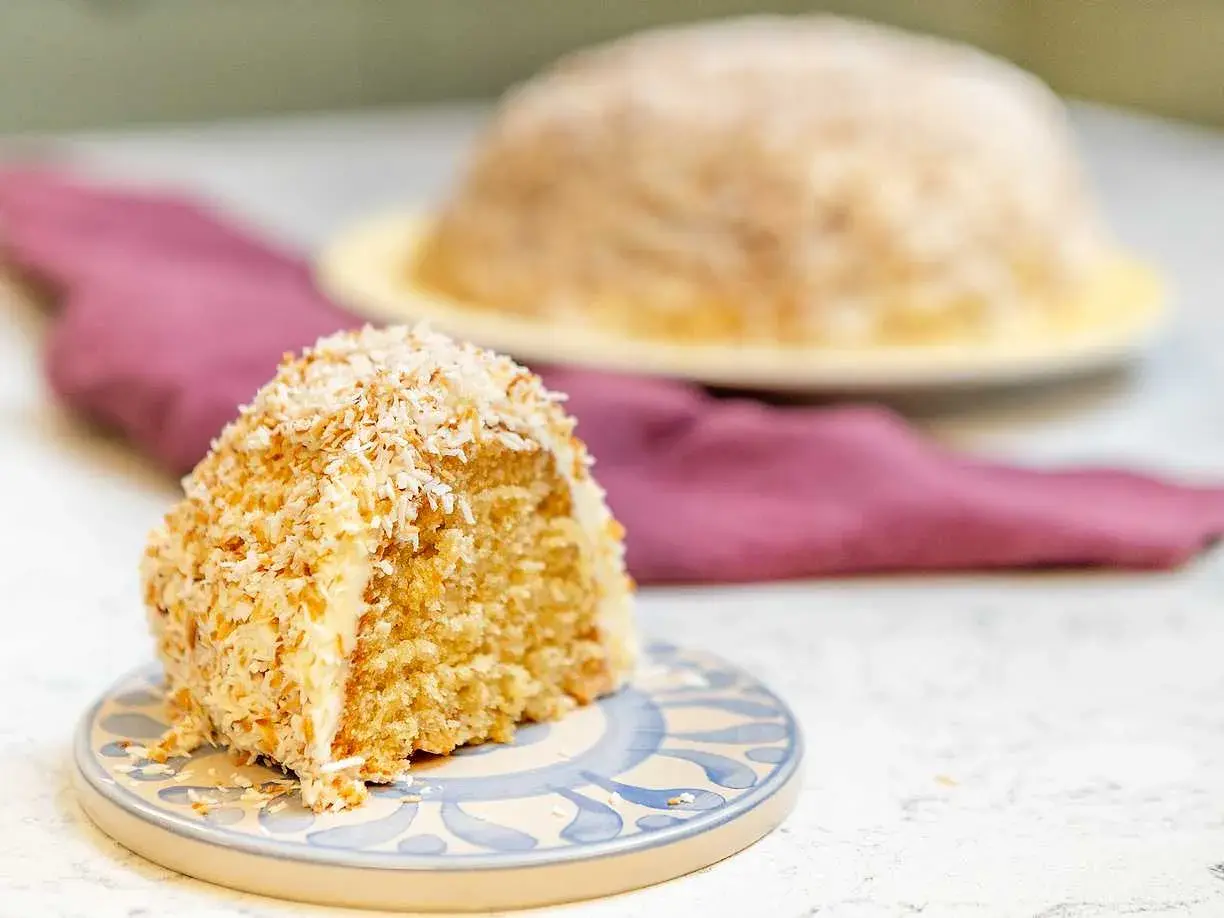 En bit sockerkaka med kokos, täckt av rostad kokosflingor. En hel kaka syns i bakgrunden.