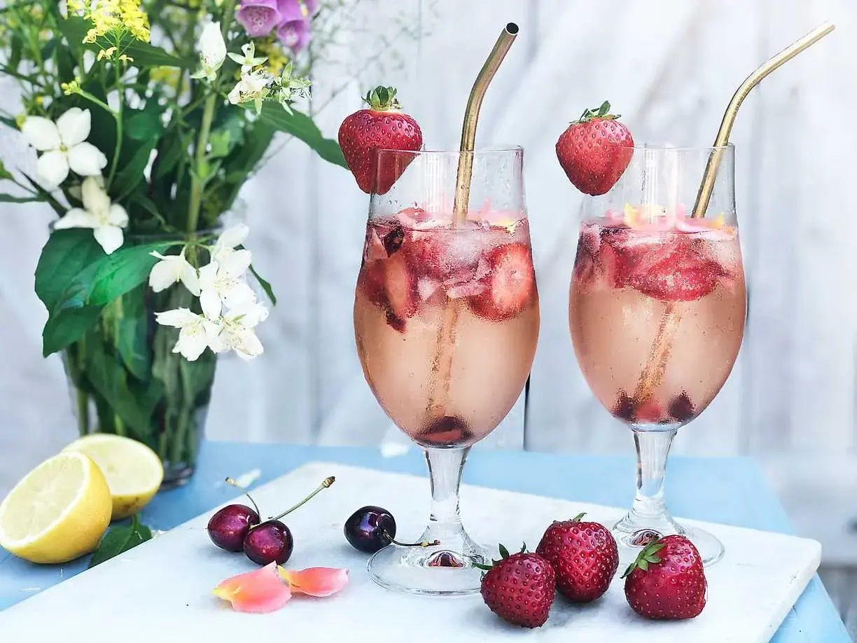 Två glas med drink med gin och prosecco, fyllda med is, jordgubbar och körsbär. Garnerade med jordgubbar och serverade med citroner och blommor.