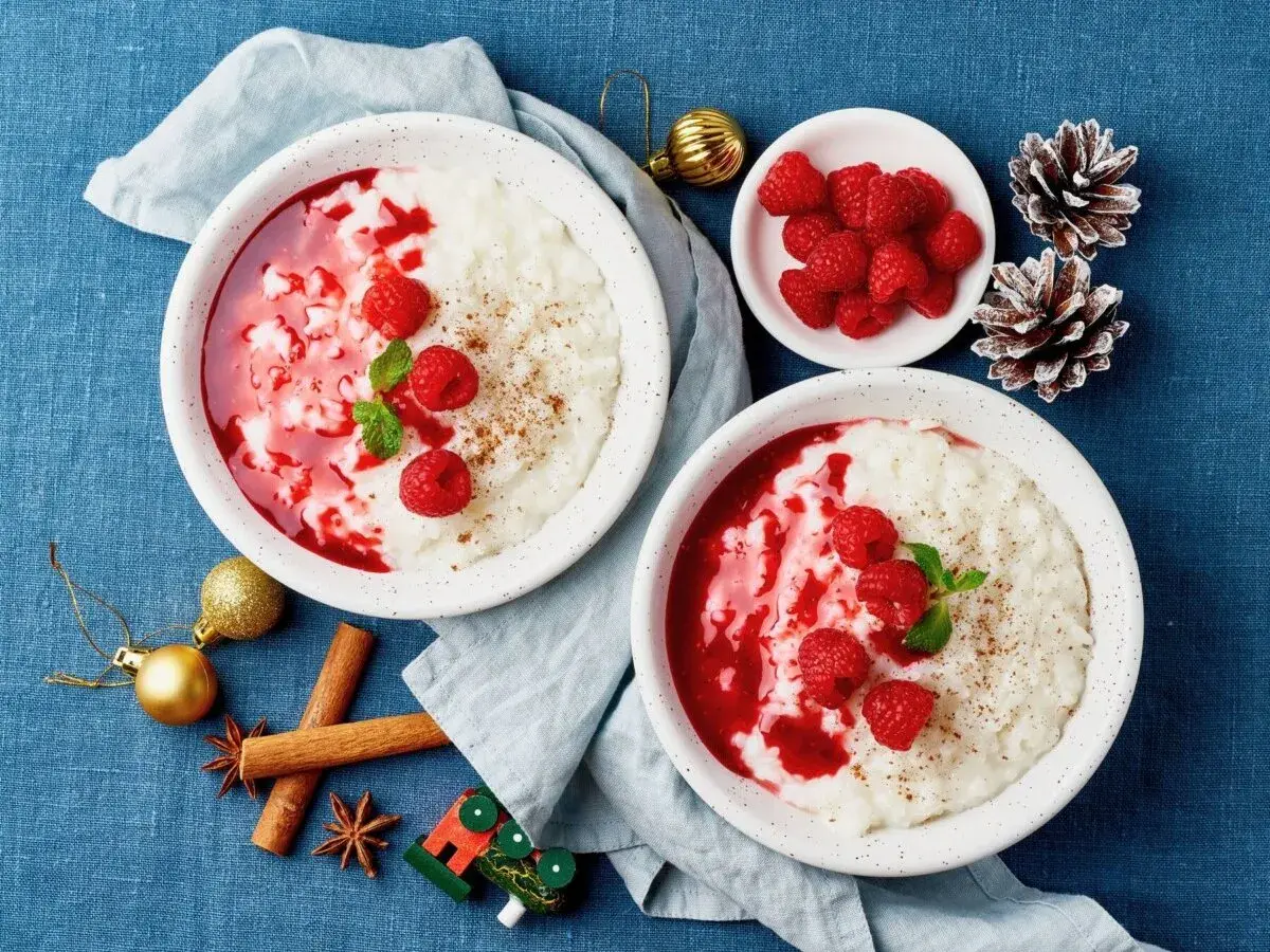 Två skålar med krämig risgrynsgröt, toppad med hallonsås, färska hallon och mynta. En julig dukning med kanelstänger och kottar. Perfekt ris ala malta!