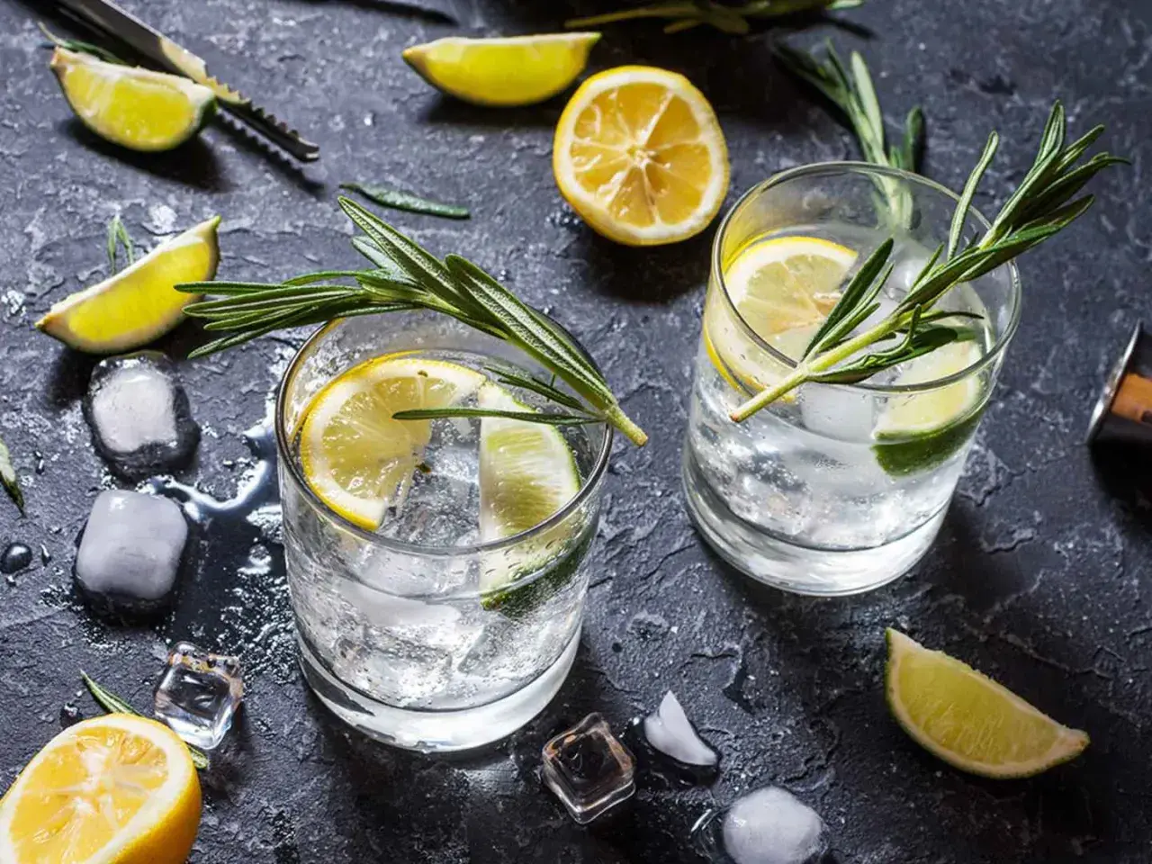 Två glas med gin och tonic, is, citron- och limeklyftor samt rosmarinkvistar på ett mörkt underlag.