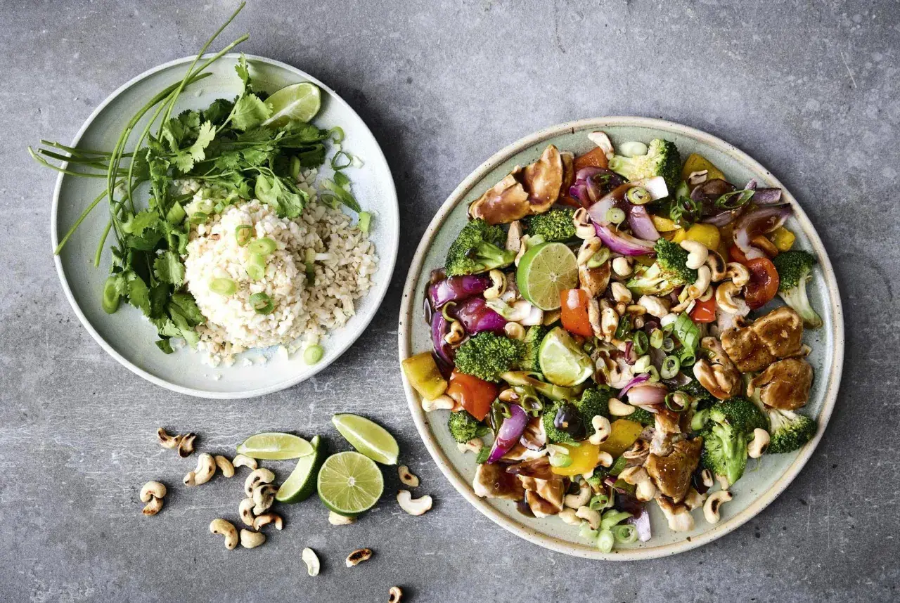 Two plates of food: one with rice, cilantro, and lime; the other with stir-fried chicken, vegetables, and cashews.