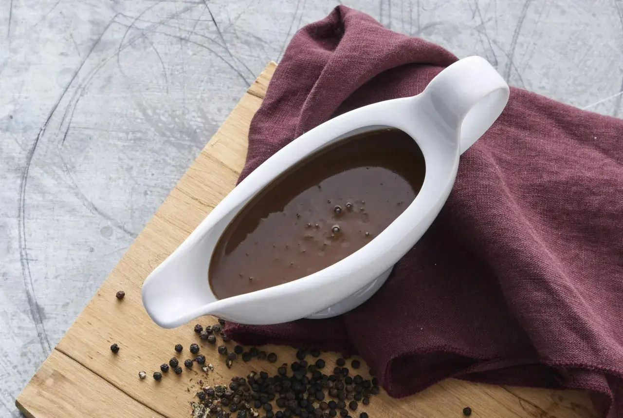 En vit såssip med mustig brunsås, redo för ett klassiskt brunsås recept. Svartpepparkorn och en vinröd duk fulländar bilden.